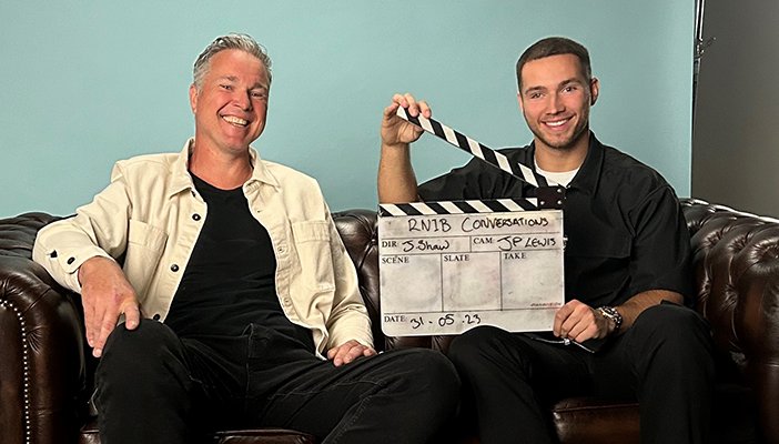 Two men sat on a sofa looking at the camera, the man on the right is holding a film clapper.