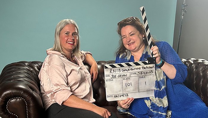 Two women are sat next to each on a sofa, the woman on the right is holding a film clapper.