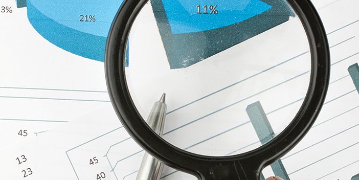 Someone holding magnifying glass over various reports with blue pie charts and bar charts.