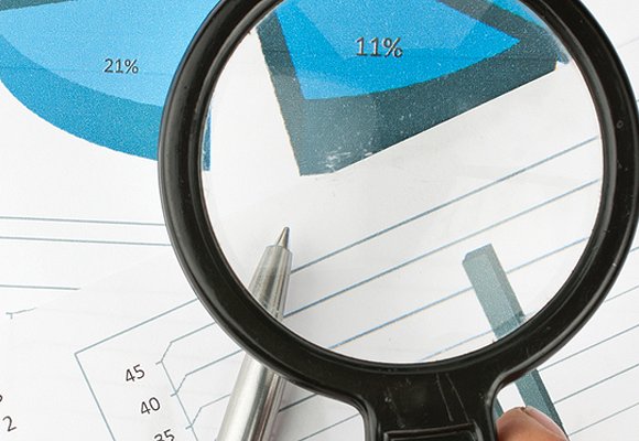 Someone holding magnifying glass over various reports with blue pie charts and bar charts.