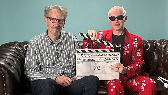 Two men are sat next to each other on a sofa, the man on the right is holding a film clapper.