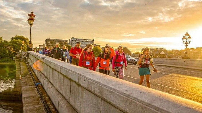 Image of Thames Path Challenge participants walking across a scenic bridge in London silhouetted by a beautiful auburn sunset.