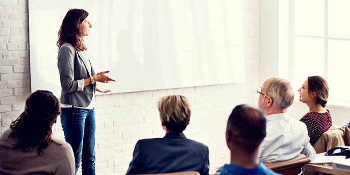 A person talking to a room full of seated people and gesturing at a whiteboard.