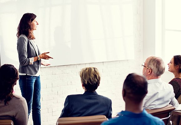 A person talking to a room full of seated people and gesturing at a whiteboard.