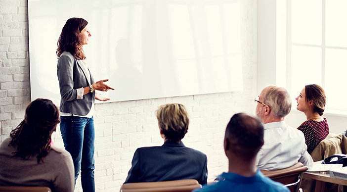 A person talking to a room full of seated people and gesturing at a whiteboard.