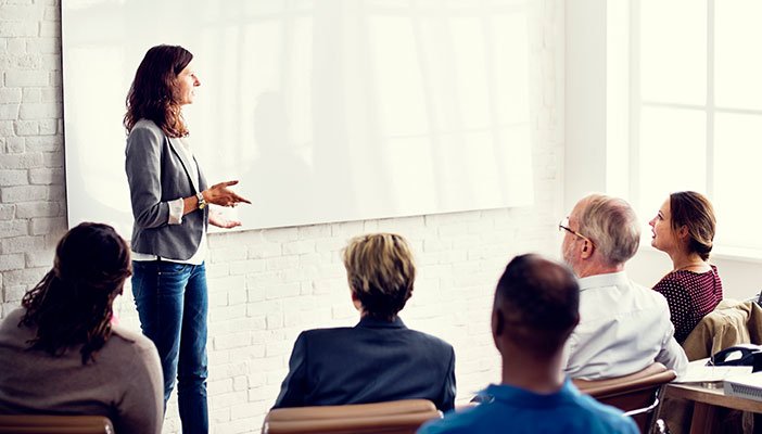 A person talking to a room full of seated people and gesturing at a whiteboard.