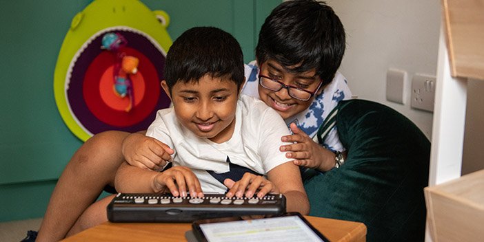 A child smiling, using a brailler with his brother smiling and sitting closely behind him looking at what he is doing.