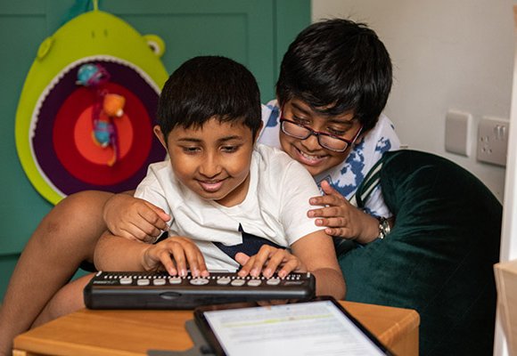 A child smiling, using a brailler with his brother smiling and sitting closely behind him looking at what he is doing.