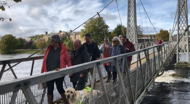 A group of people and a guide dog cross a metal bridge over a large body of water.