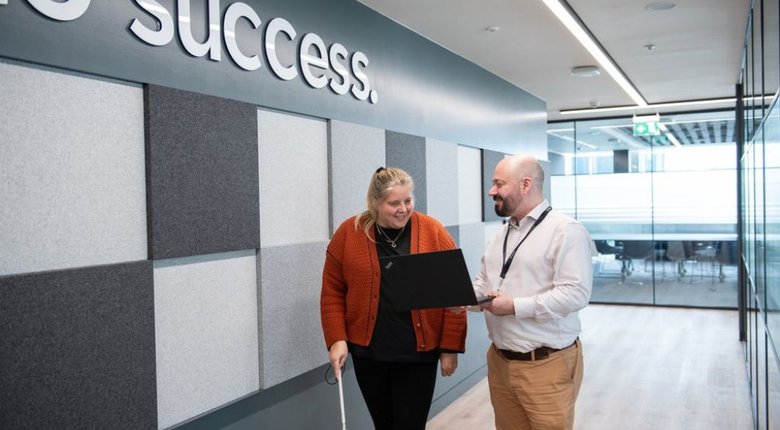 A woman using a cane stands next to a man holding a laptop in an office corridor. The man is showing the woman something on the laptop screen.