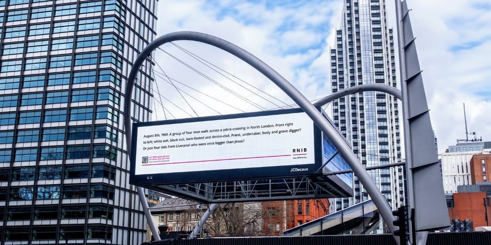 A digital billboard in between two glass skyscrapers in London. Black text on a white background on the billboard reads 'August 8th, 1969. A group of four men walk across a zebra crossing in North London. From right to left, white suit, black suit, bare-footed and denim-clad. Priest, undertaker, body and grave differ? Or just four lads from Liverpool who were once bigger than Jesus?'