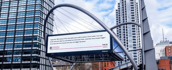 A digital billboard in between two glass skyscrapers in London. Black text on a white background on the billboard reads 'August 8th, 1969. A group of four men walk across a zebra crossing in North London. From right to left, white suit, black suit, bare-footed and denim-clad. Priest, undertaker, body and grave differ? Or just four lads from Liverpool who were once bigger than Jesus?'