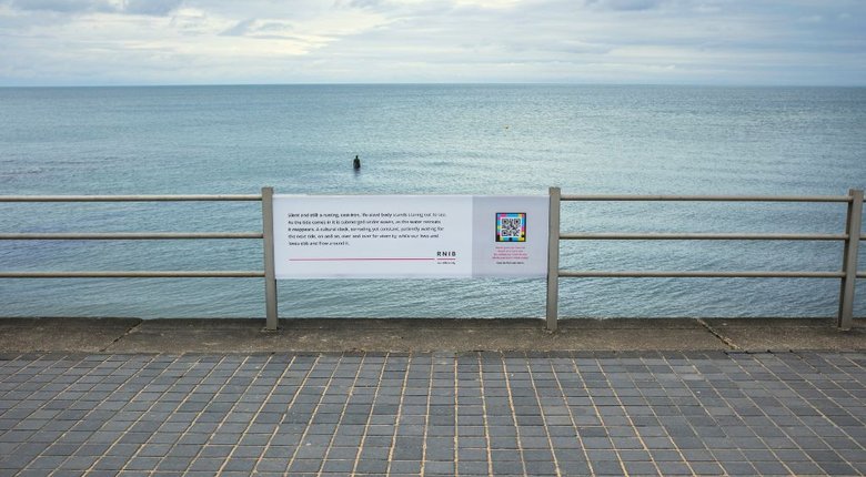 In the distance a lone statue figure is stood in the sea up to waist height. In the foreground is a white sign on a metal railing. The text on the sign reads 'silent and still: a rusting, cast-iron, life-size body stands staring out to sea. As the tide comes in it is submerged under waves, as the water retreats it reappears. A cultural clock, corroding yet constant, patiently waiting for the next tide, on and on, over and over for eternity, while our lives and loves ebb and flow around it.'