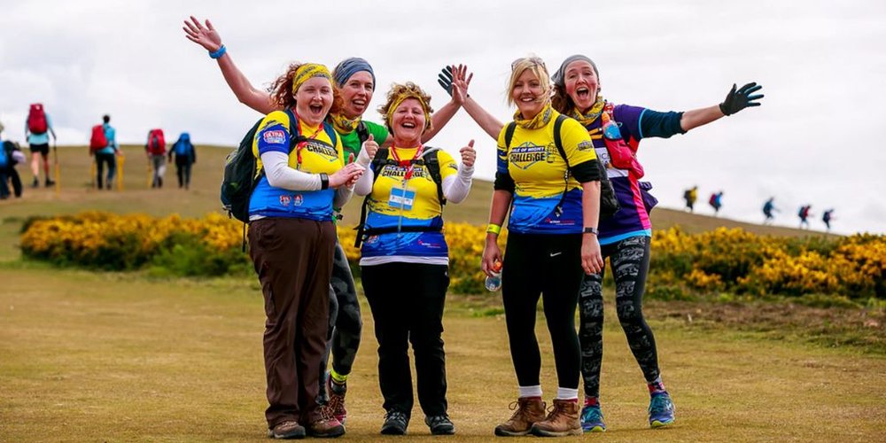 A group of five white women taking part in an Ultra Challenge. They are all wearing activewear, walking boots and backpacks stand on a grass hill grinning at the camera.