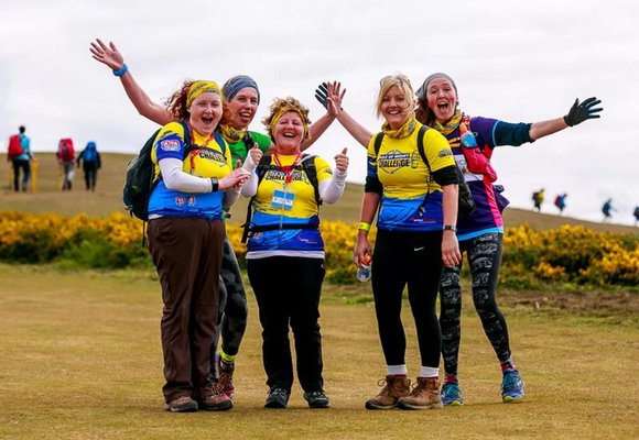 A group of five white women taking part in an Ultra Challenge. They are all wearing activewear, walking boots and backpacks stand on a grass hill grinning at the camera.