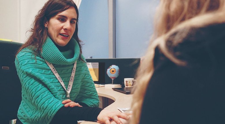 A white woman with shoulder length brown hair, wearing a turquoise high neck knitted jumper, sits at a desk and talks to someone. Only the back of the person she is talking to is visible.