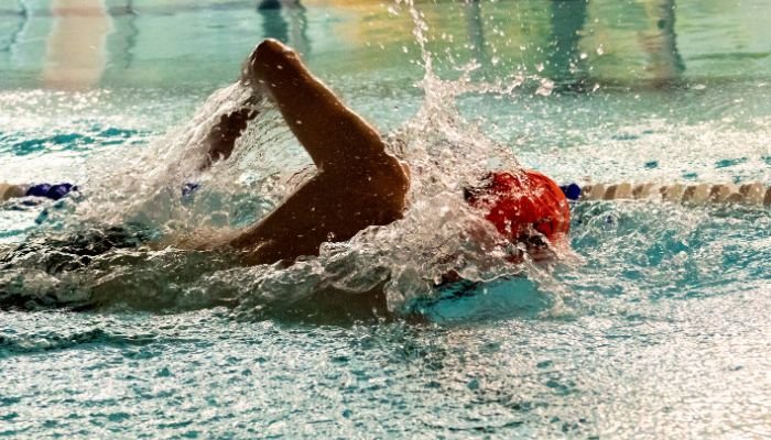 Side view of person swimming front crawl in an indoor swimming pool, wearing an orange swim cap.
