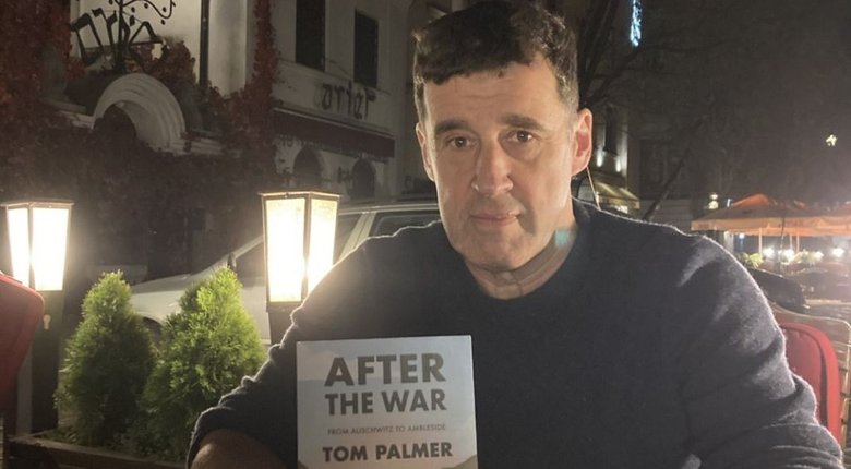 Tom Palmer, an older white man with short dark hair, looks directly at the camera. He is seated and holding a copy of his book 'After the War.' There are large lit garden lights and bushes in the background.