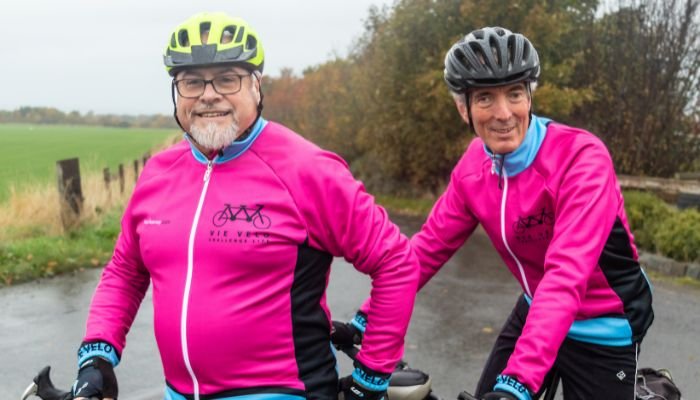 Two men wearing cycling helmets and pink and blue cycling zip up tops, both riding a tandem bicycle.
