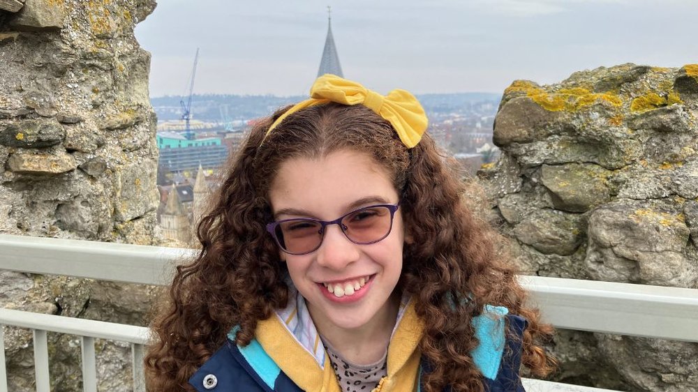 Rosie Davison, smiles widely at the camera. She is a 10 year old girl with curly brow hair with a big yellow bow on top of her head. She's wearing glasses with purple frames and a blue waterproof coat. Behind her is a metal railing and stone wall, and in the background is a cityscape.