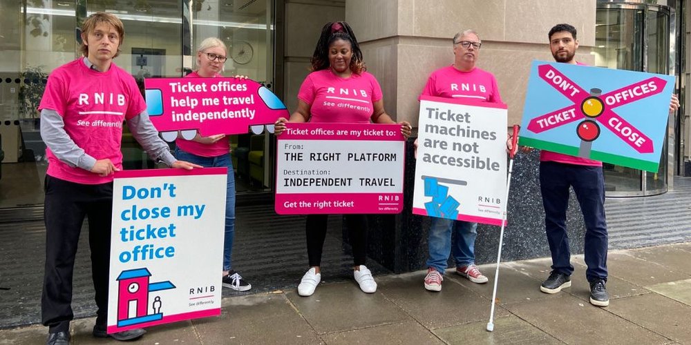 Five people wearing pink RNIB t-shirts and holding signs protesting railway stations ticket office closures, stand outside the Department for Transport.
