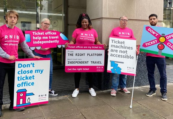 Five people wearing pink RNIB t-shirts and holding signs protesting railway stations ticket office closures, stand outside the Department for Transport.