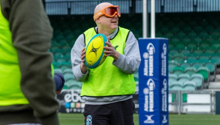 A man playing visually impaired rugby, wearing orange goggles and a grey sweatshirt with a fluorescent yellow tabard over it. Holding a rugby ball.