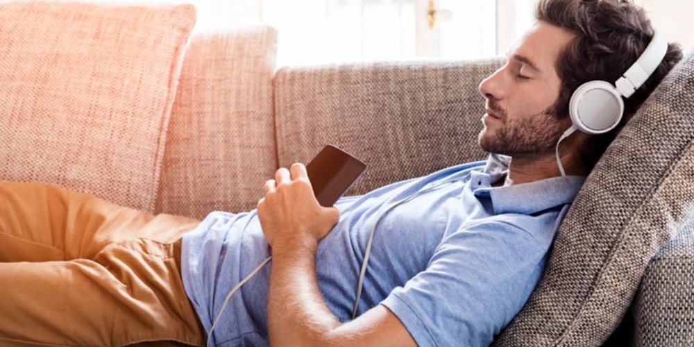 A man lying on a sofa, wearing headphones, listening to audio from an IPhone