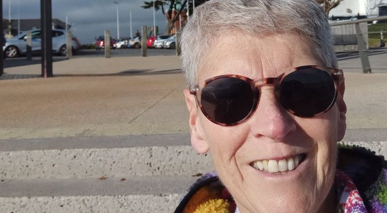 Una Mulgrew, an older white woman with very short grey hair, wearing sunglasses. She is on the right hand side of the image, smiling directly at the camera as she takes a selfie. In the left hand side and background of the image is a car park that's out of focus.