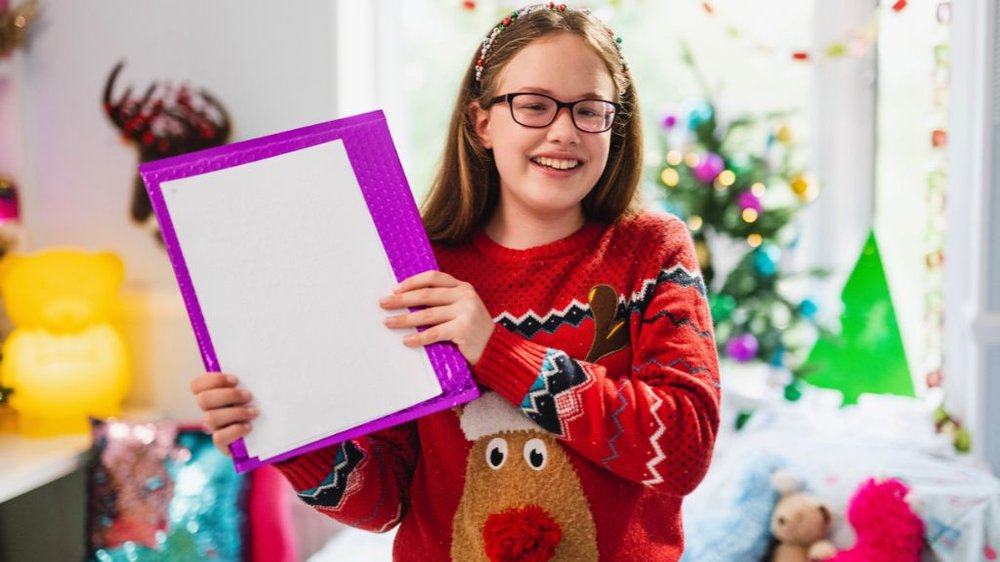 An image of our chief Joy Maker Keira, stood smiling in front of Christmas decorations whilst wearing a red festive jumper and proudly holding up her accessible Letter from Santa.