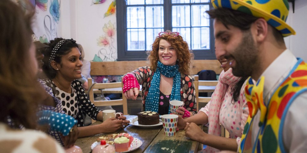 A group of people wearing dotty clothes and drinking tea sat at a table with a variety of cakes on it.