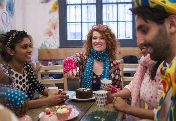 A group of people wearing dotty clothes and drinking tea sat at a table with a variety of cakes on it.