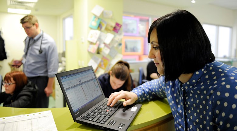 A woman at a busy hospital reception desk working on a laptop.