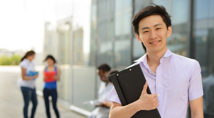 A student holding a clipboard in front of a college building with two students in the background.