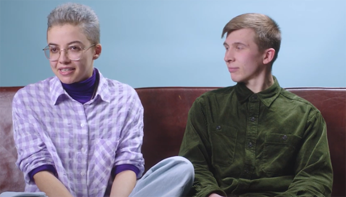 Two people sitting on a sofa, the person on the left is wearing glasses and looking at the camera.