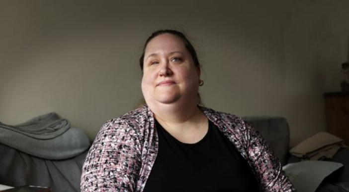 Hazel Hyland sits on her sofa which is upholstered in grey fabric she is wearing a black top with a black and white shirt over the top.