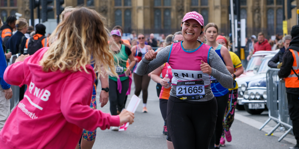 Aidana smiling whilst running past the RNIB cheer point at the London Landmarks Half Marathon. On the left of the image you can see the back of someone wearing a pink RNIB hoodie, cheering Aidana on. Running behind Aidana are other people taking part in the London Landmarks Half Marathon, but they are not in focus.