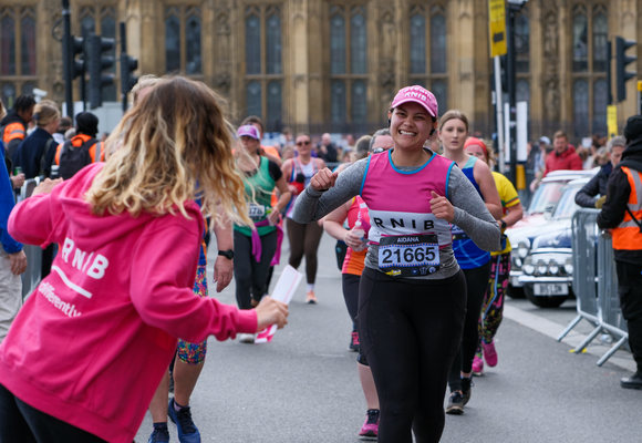 Aidana smiling whilst running past the RNIB cheer point at the London Landmarks Half Marathon. On the left of the image you can see the back of someone wearing a pink RNIB hoodie, cheering Aidana on. Running behind Aidana are other people taking part in the London Landmarks Half Marathon, but they are not in focus.