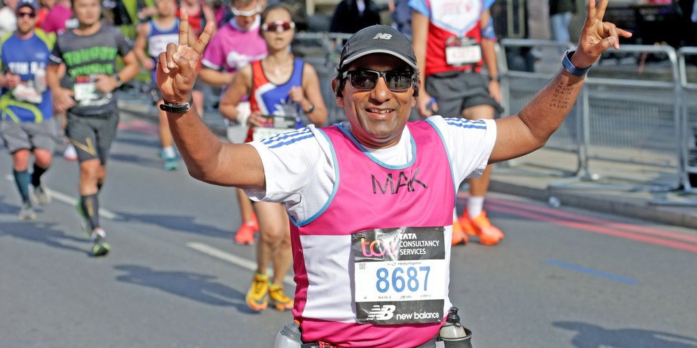Mak, a man wearing black shorts, a black baseball cap, sunglasses, a white t-shirt, a pink running vest and a running number, smiles at the camera with his arms in the air as he gives a peace sign with both hands. He is running the London 10k. There are more runners in the background but Mak is the only one in focus. They are running on the road.
