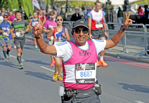 Mak, a man wearing black shorts, a black baseball cap, sunglasses, a white t-shirt, a pink running vest and a running number, smiles at the camera with his arms in the air as he gives a peace sign with both hands. He is running the London 10k. There are more runners in the background but Mak is the only one in focus. They are running on the road.