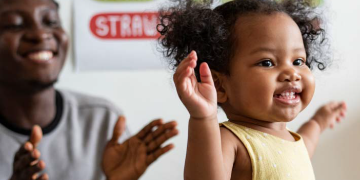 A parent claps and smiles as a toddler walks.