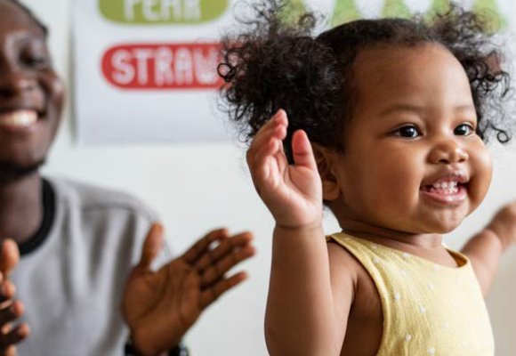 A parent claps and smiles as a toddler walks.
