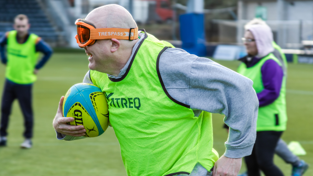A man holding a rugby ball with eye shades on