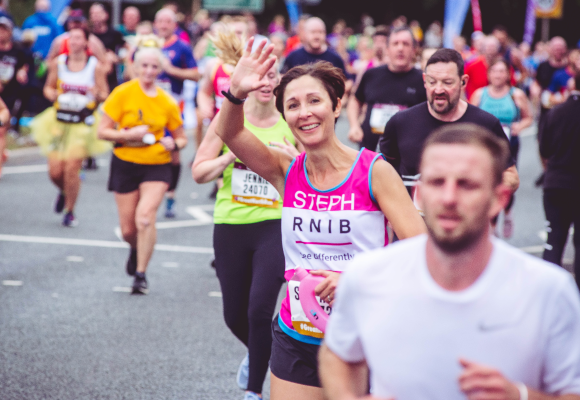A large group of runners. One is wearing a pink RNIB shirt and waving to the camera.