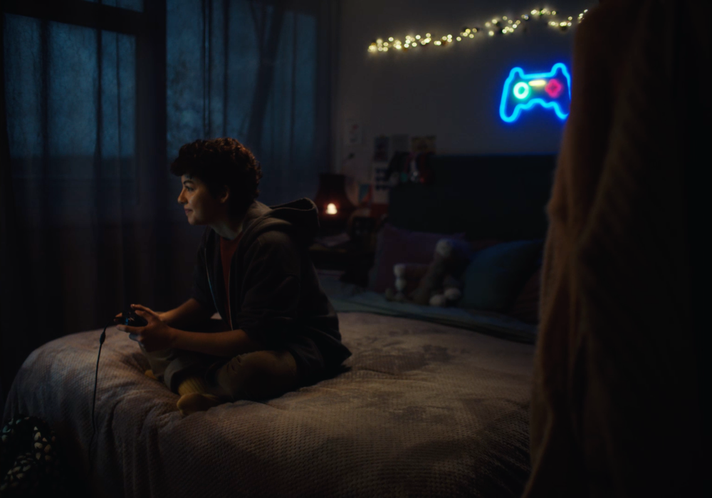 Ava, a young woman with short hair sits on the edge of their bed, playing a video game with a controller. The room is dimly lit, with soft lighting from a neon game controller sign on the wall and string lights above the bed. The person appears focused on the game. Note for the web team- this image is already on our website.