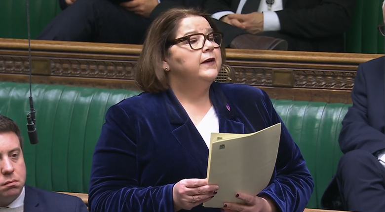 Julie Minns MP stood in the House of Commons delivering a speech presenting the Elections (Accessibility for Blind Voters) Bill. Julie is a white woman with dark brown hair cut into a bob. She's wearing black rimmed glasses and a dark blue velvet blazer over a white t-shirt.