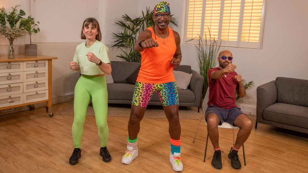From left: Sarah (wearing green) and Mr Motivator (wearing orange vest and multi-coloured bandana and shorts) are stood up, doing a punching forwards exercise. Yahya is seated on a chair, doing the same thing, in a living room setting.
